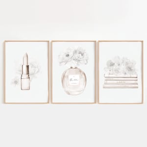 Puede incluir: Tres impresiones en acuarela enmarcadas. La impresión de la izquierda presenta una barra de labios con un fondo floral. La impresión central muestra un frasco de perfume con la palabra "Belle" y flores. La impresión de la derecha muestra una pila de libros con flores.
