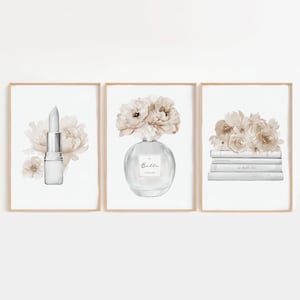 Op de afbeelding: Drie ingelijste aquarelprints met illustraties met een schoonheidsthema. Eén print toont een lippenstift, een andere een parfumfles en de derde een stapel boeken, allemaal met bloemaccenten in neutrale tinten.