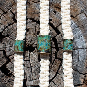 May include: Three cream-colored macrame cords with ceramic beads. The beads have a turquoise and brown swirl pattern. The cords are displayed against a weathered, textured wood background.