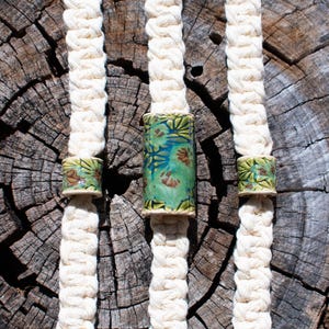 May include: Three cream-colored, braided cords with ceramic beads. The beads have a green, blue, and brown floral design. The cords are displayed against a textured, weathered wood background.