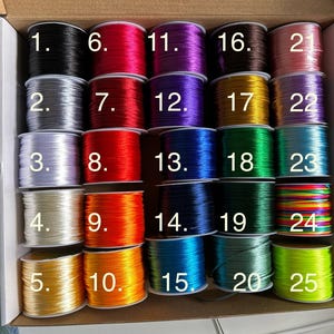 May include: A variety of colorful spools of cord, numbered 1 through 25, are arranged in a cardboard box. The cords are in various colors, including black, silver, red, orange, yellow, green, blue, and purple. The image is well-lit, showcasing the vibrant colors of the cords.