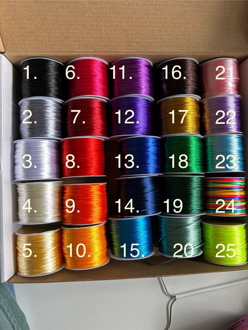 May include: A variety of colorful spools of cord, numbered 1 through 25, are arranged in a cardboard box. The cords are in various colors, including black, silver, red, orange, yellow, green, blue, and purple. The image is well-lit, showcasing the vibrant colors of the cords.