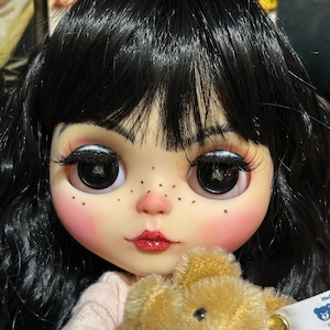 May include: A close-up of a doll with black hair, large eyes, and rosy cheeks. The doll is holding a small, tan teddy bear. The doll's lips are painted red, and it has freckles. The background is blurred.