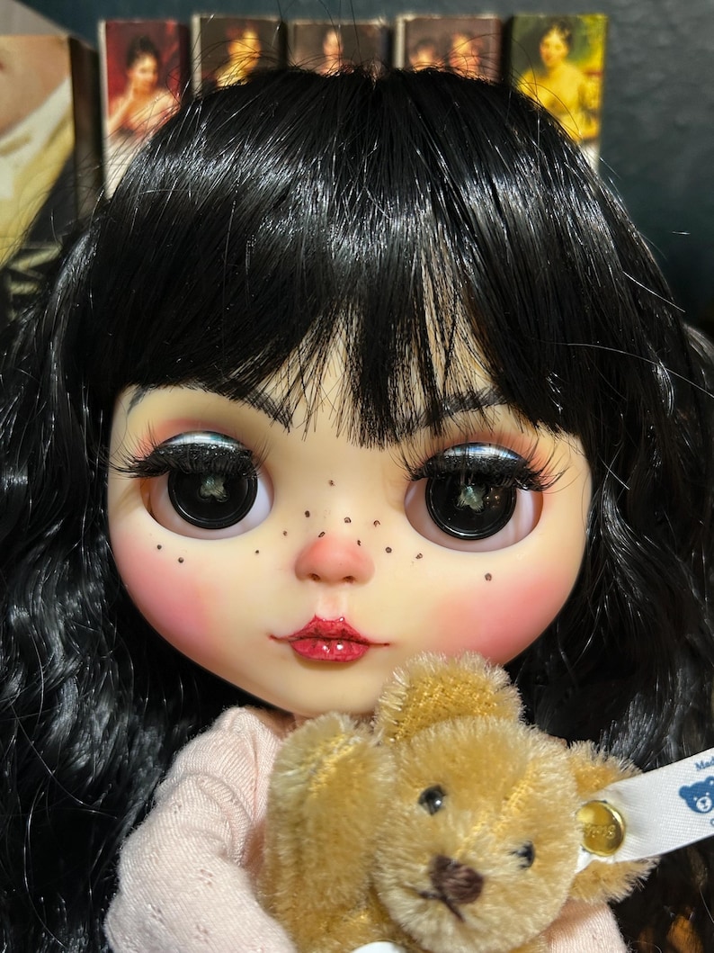 May include: A close-up of a doll with black hair, large eyes, and rosy cheeks. The doll is holding a small, tan teddy bear. The doll's lips are painted red, and it has freckles. The background is blurred.