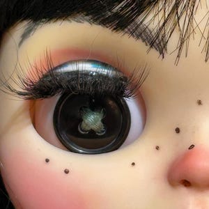 May include: Close-up of a doll's face with a button eye. The eye is a large, black button with a cross-stitch design. The doll has long, black eyelashes, black hair, and freckles. The skin is pale with rosy cheeks and red lips.