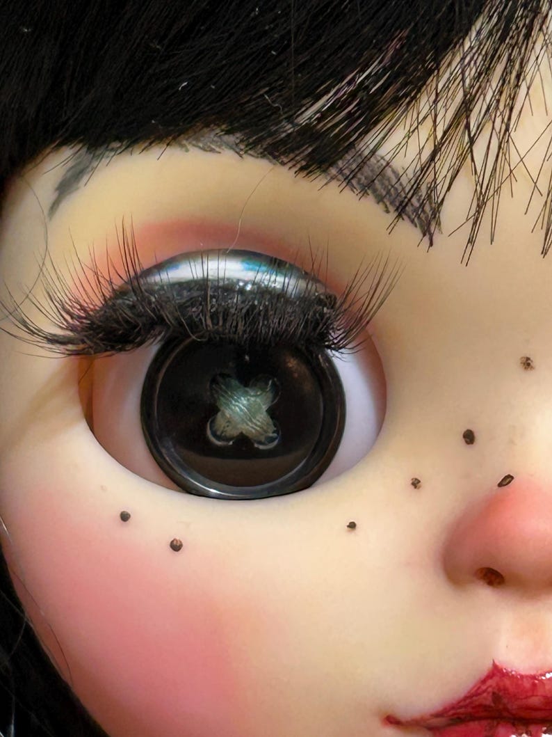 May include: Close-up of a doll's face with a button eye. The eye is a large, black button with a cross-stitch design. The doll has long, black eyelashes, black hair, and freckles. The skin is pale with rosy cheeks and red lips.