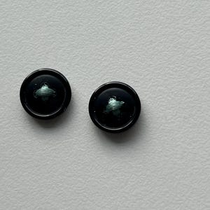 May include: Two black buttons with a small, stylized green design in the center. The buttons are round and have a glossy finish. They are set against a plain, light-colored background.