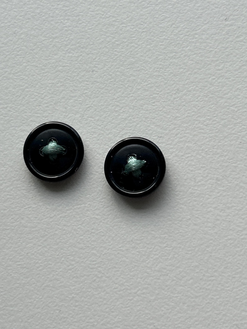May include: Two black buttons with a small, stylized green design in the center. The buttons are round and have a glossy finish. They are set against a plain, light-colored background.