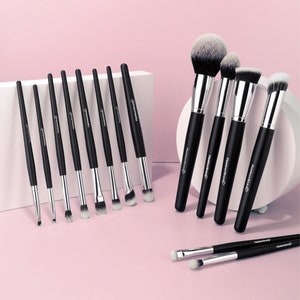 May include: A set of black makeup brushes with silver ferrules and white bristles. The brushes are arranged on a pink surface. The handles have the text "Flawless Beauty" printed on them. The set includes various brush sizes and shapes.