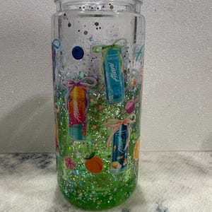 May include: A clear glass tumbler with a lid, filled with green liquid and multi-colored glitter. The exterior features various printed graphics including Alani energy drink cans tied with bows, a blueberry, a peach, and small star shapes.