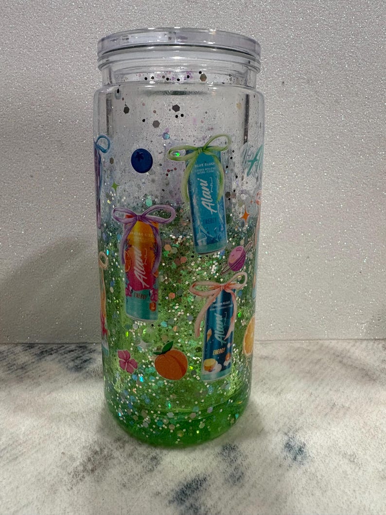 May include: A clear glass tumbler with a lid, filled with green liquid and multi-colored glitter. The exterior features various printed graphics including Alani energy drink cans tied with bows, a blueberry, a peach, and small star shapes.
