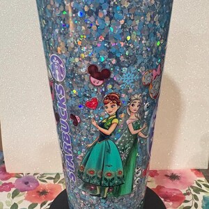 May include: A clear plastic Starbucks tumbler filled with blue and silver holographic glitter. The exterior features printed images of two animated characters in green and teal dresses, a red heart, Mickey Mouse ears, and the Starbucks logo text.