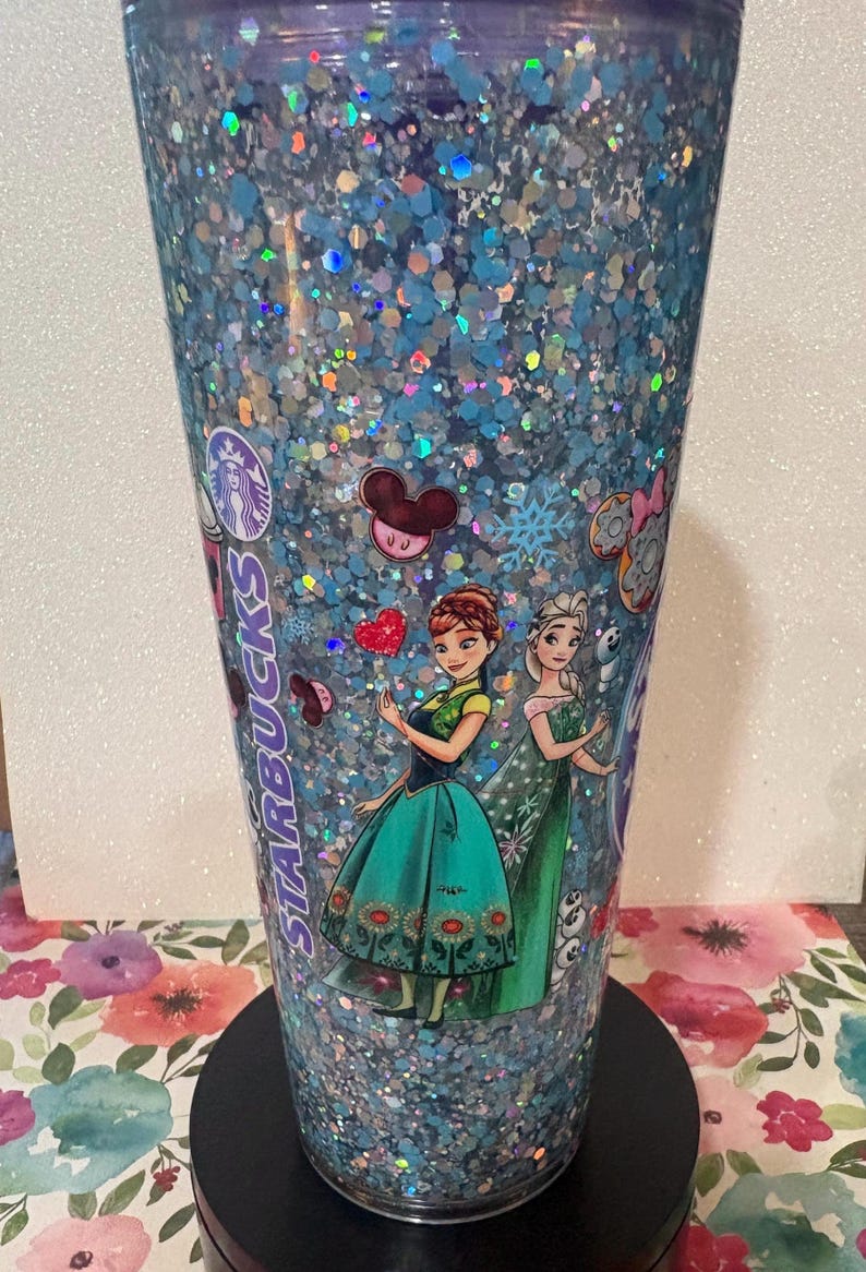May include: A clear plastic Starbucks tumbler filled with blue and silver holographic glitter. The exterior features printed images of two animated characters in green and teal dresses, a red heart, Mickey Mouse ears, and the Starbucks logo text.