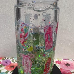 May include: A clear glass tumbler with a lid, filled with green liquid and multi-colored glitter. The exterior features various Alani energy drink can graphics, each tied with a small bow, along with small lime slice and star illustrations.