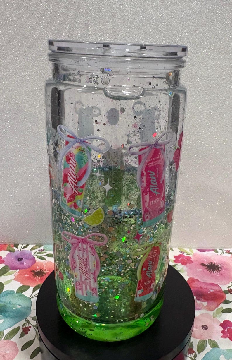 May include: A clear glass tumbler with a lid, filled with green liquid and multi-colored glitter. The exterior features various Alani energy drink can graphics, each tied with a small bow, along with small lime slice and star illustrations.