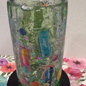 May include: A clear tumbler filled with green liquid and iridescent glitter, featuring various Alani energy drink can graphics, small fruit illustrations, and decorative bows printed on the exterior, topped with a clear plastic lid.