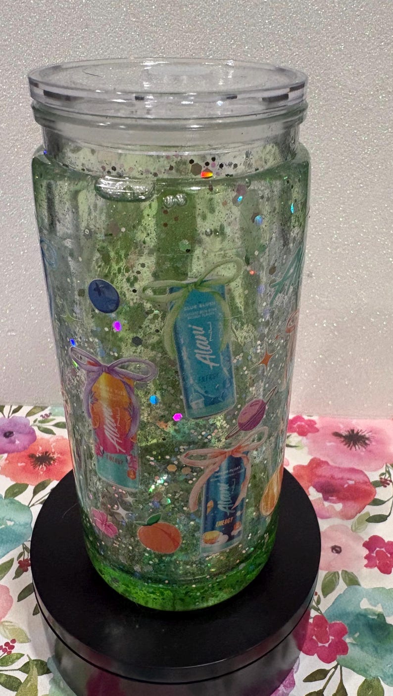 May include: A clear tumbler filled with green liquid and iridescent glitter, featuring various Alani energy drink can graphics, small fruit illustrations, and decorative bows printed on the exterior, topped with a clear plastic lid.