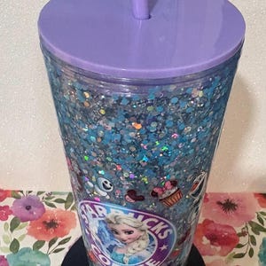 May include: A clear plastic tumbler filled with blue and silver holographic glitter, featuring a purple lid and matching straw. The exterior displays a circular logo with the text Starbucks Coffee and an illustration of Elsa, alongside small character icons.