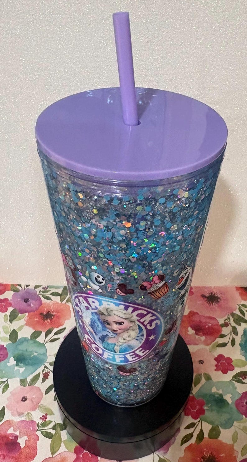 May include: A clear plastic tumbler filled with blue and silver holographic glitter, featuring a purple lid and matching straw. The exterior displays a circular logo with the text Starbucks Coffee and an illustration of Elsa, alongside small character icons.