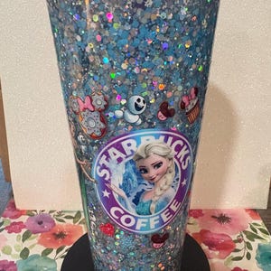 May include: A clear plastic tumbler with a purple lid, filled with blue and silver holographic glitter. The exterior features a circular Starbucks logo with Elsa from Frozen in the centre, surrounded by small decals of Olaf, doughnuts, and cupcakes.