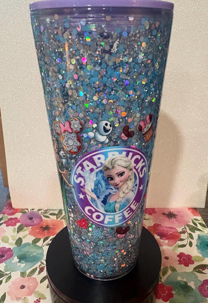 May include: A clear plastic tumbler with a purple lid, filled with blue and silver holographic glitter. The exterior features a circular Starbucks logo with Elsa from Frozen in the centre, surrounded by small decals of Olaf, doughnuts, and cupcakes.