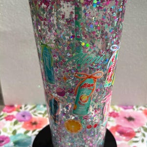 May include: A clear plastic tumbler with a mint green lid and straw, filled with iridescent pink and silver glitter. The exterior features various Alani Nu energy drink can graphics, orange slices, cherries, and small planet illustrations.