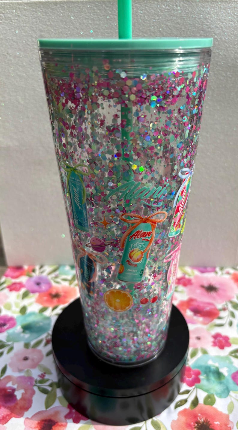 May include: A clear plastic tumbler with a mint green lid and straw, filled with iridescent pink and silver glitter. The exterior features various Alani Nu energy drink can graphics, orange slices, cherries, and small planet illustrations.