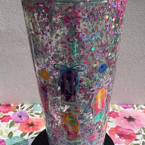 May include: A clear plastic tumbler with a mint green lid and straw, filled with multi-colored pink, blue, and silver glitter. The exterior features printed graphics of various Alani energy drink cans, each tied with a decorative bow.