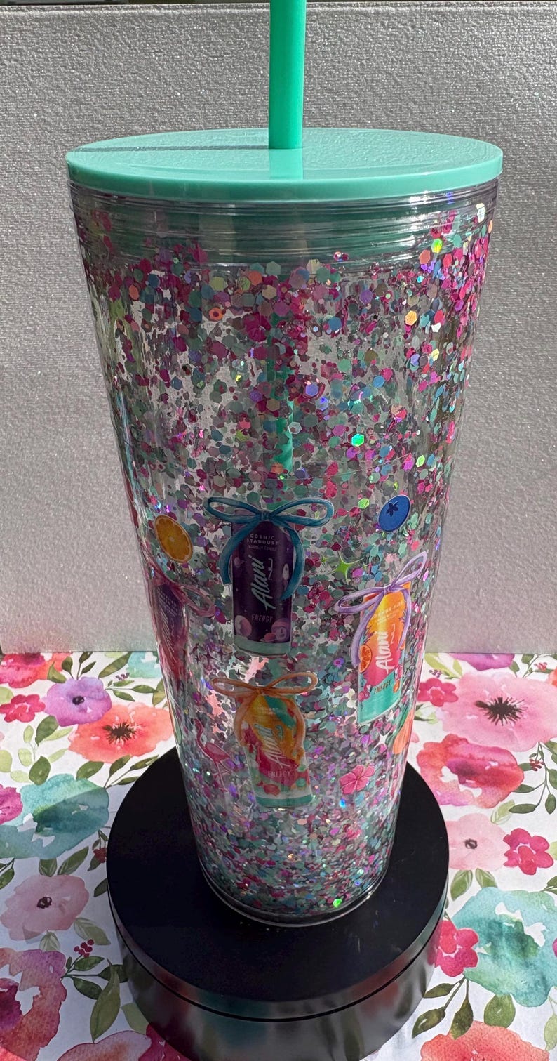 May include: A clear plastic tumbler with a mint green lid and straw, filled with multi-colored pink, blue, and silver glitter. The exterior features printed graphics of various Alani energy drink cans, each tied with a decorative bow.