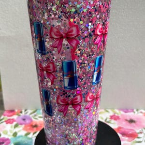 May include: A clear, pink glitter tumbler with a matching pink lid and straw. The tumbler is decorated with pink bows and blue Red Bull can decals. The tumbler sits on a black base and is filled with pink and iridescent glitter.