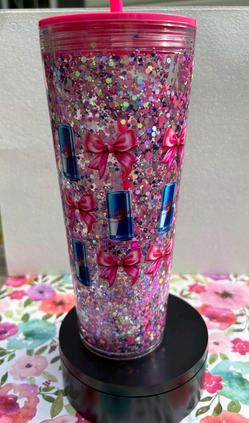 May include: A clear, pink glitter tumbler with a matching pink lid and straw. The tumbler is decorated with pink bows and blue Red Bull can decals. The tumbler sits on a black base and is filled with pink and iridescent glitter.