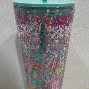 May include: A clear plastic tumbler with a teal lid and straw, filled with pink and silver glitter. The exterior features printed graphics of Alani energy drink cans tied with bows, along with small illustrations of peach, lime, and orange fruit slices.