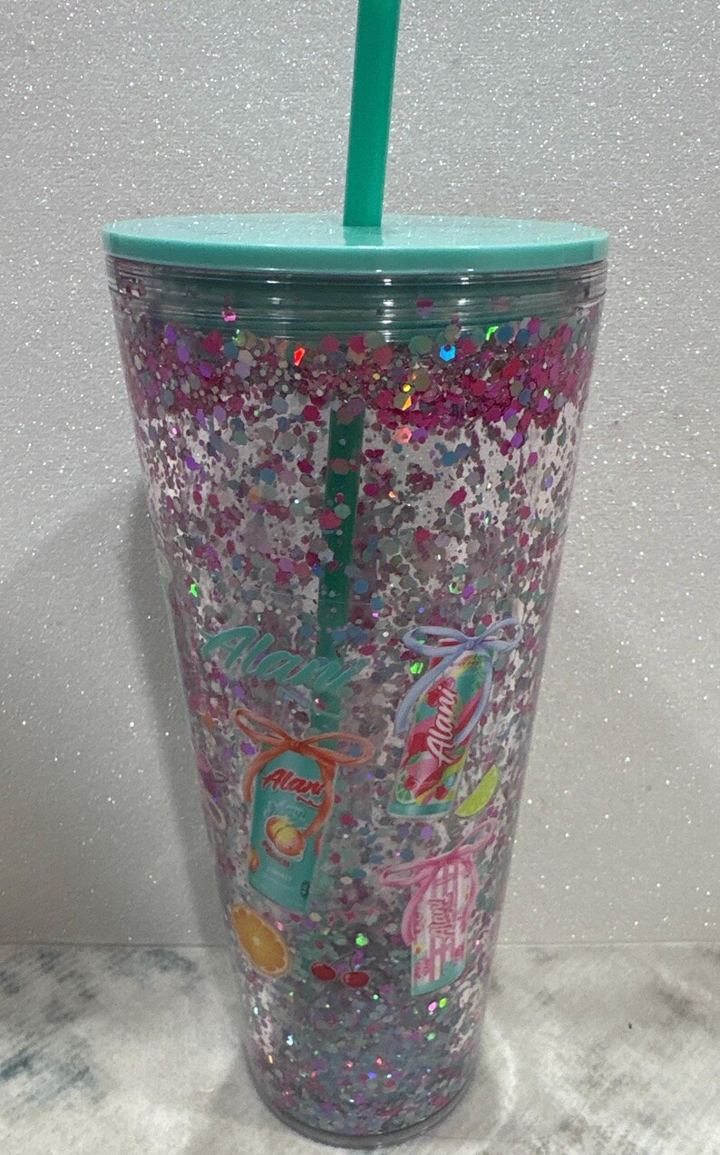 May include: A clear plastic tumbler with a teal lid and straw, filled with pink and silver glitter. The exterior features printed graphics of Alani energy drink cans tied with bows, along with small illustrations of peach, lime, and orange fruit slices.