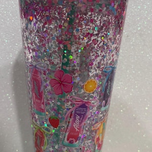 May include: A clear plastic tumbler with a mint green lid and straw, filled with pink and silver holographic glitter. The exterior features printed graphics of Alani energy drink cans, hibiscus flowers, citrus slices, strawberries, and a flamingo.