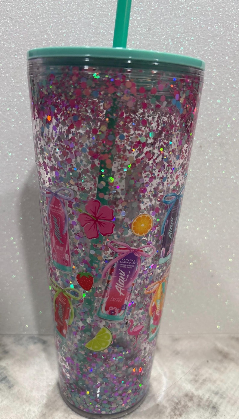 May include: A clear plastic tumbler with a mint green lid and straw, filled with pink and silver holographic glitter. The exterior features printed graphics of Alani energy drink cans, hibiscus flowers, citrus slices, strawberries, and a flamingo.