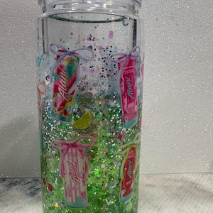 May include: A clear double-walled tumbler filled with green liquid and multi-colored glitter. The exterior features printed graphics of Alani energy drink cans tied with bows, lime slices, and cherries. The tumbler includes a clear plastic lid.