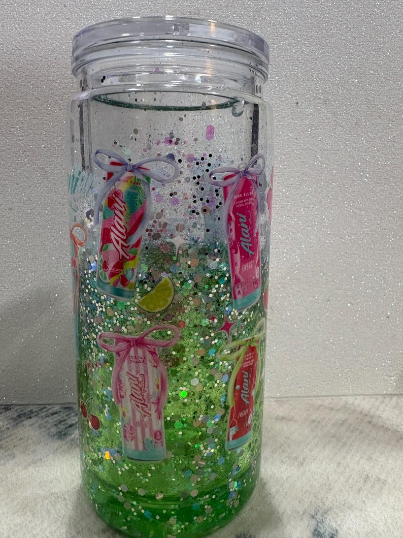 May include: A clear double-walled tumbler filled with green liquid and multi-colored glitter. The exterior features printed graphics of Alani energy drink cans tied with bows, lime slices, and cherries. The tumbler includes a clear plastic lid.