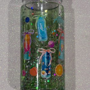 May include: A clear glass tumbler filled with green liquid and iridescent glitter. The exterior features various stickers of Alani energy drink cans, each tied with a decorative bow, alongside small icons of a peach, an orange, and a planet.