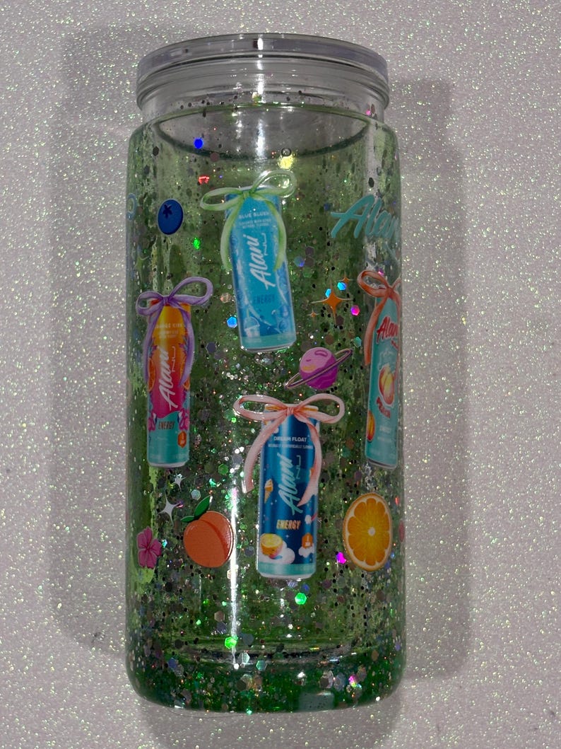 May include: A clear glass tumbler filled with green liquid and iridescent glitter. The exterior features various stickers of Alani energy drink cans, each tied with a decorative bow, alongside small icons of a peach, an orange, and a planet.