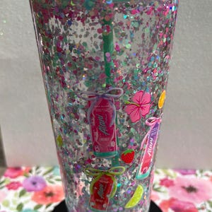 May include: A clear double-walled tumbler filled with pink, blue, and silver glitter, featuring a mint green lid and straw. The exterior is decorated with small illustrations of Alani energy drink cans, hibiscus flowers, strawberries, and lime slices.