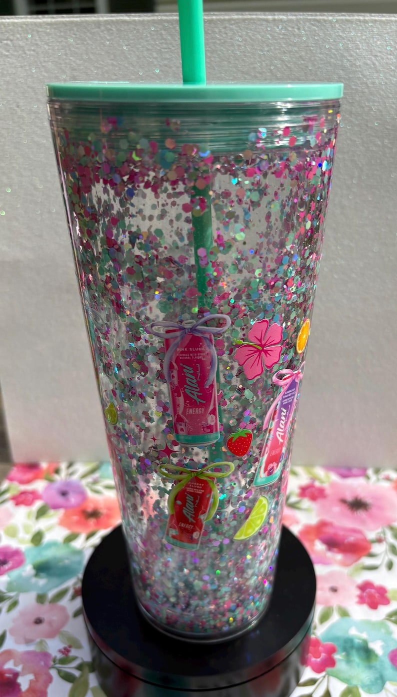 May include: A clear double-walled tumbler filled with pink, blue, and silver glitter, featuring a mint green lid and straw. The exterior is decorated with small illustrations of Alani energy drink cans, hibiscus flowers, strawberries, and lime slices.