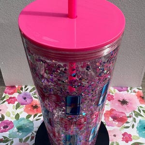 May include: A clear tumbler with a pink lid and straw, filled with pink glitter and gift box designs. The tumbler sits on a black base, with a floral patterned background.