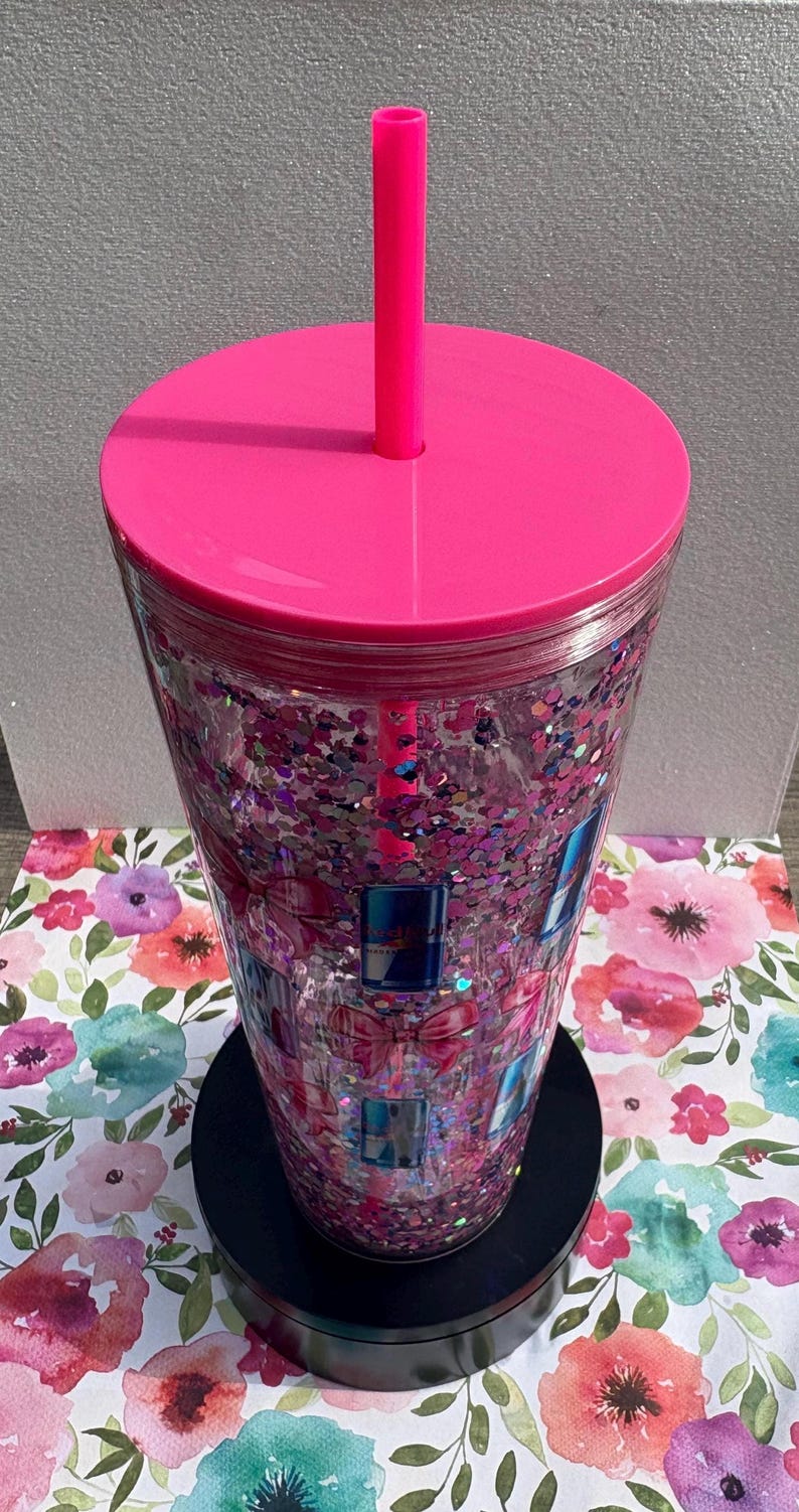 May include: A clear tumbler with a pink lid and straw, filled with pink glitter and gift box designs. The tumbler sits on a black base, with a floral patterned background.