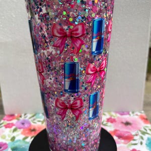 May include: A clear tumbler filled with pink glitter and iridescent confetti. The tumbler is decorated with pink bows and blue Red Bull cans. The tumbler sits on a black base and is surrounded by a floral pattern.