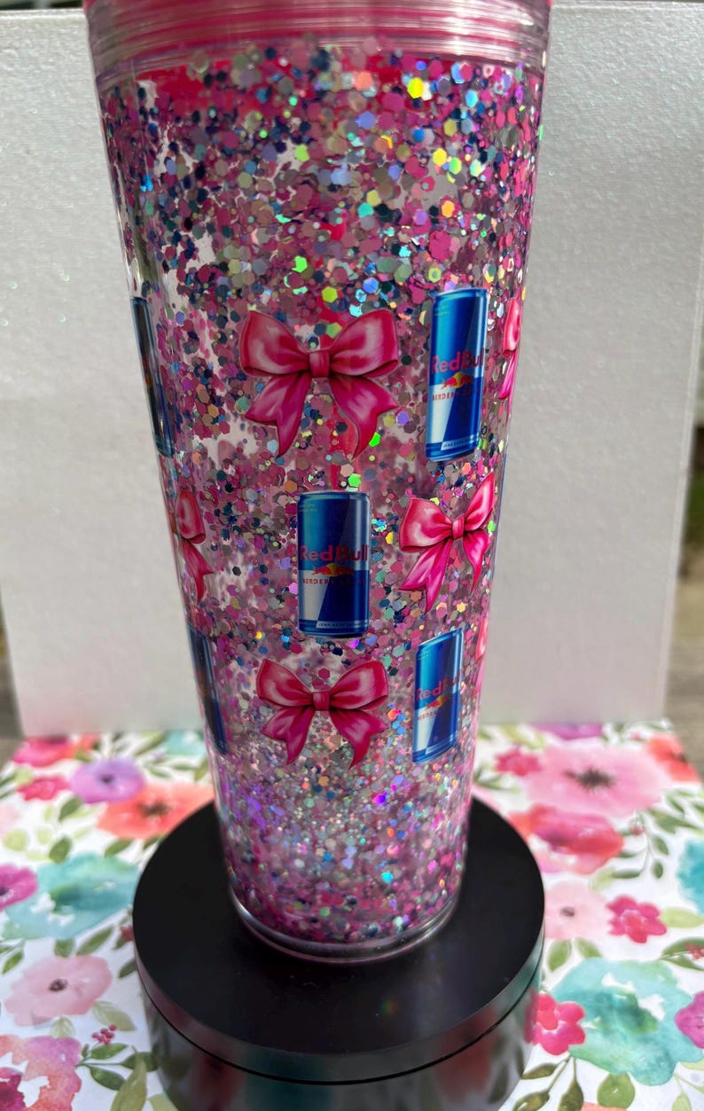 May include: A clear tumbler filled with pink glitter and iridescent confetti. The tumbler is decorated with pink bows and blue Red Bull cans. The tumbler sits on a black base and is surrounded by a floral pattern.