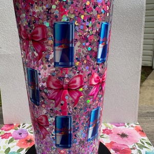 May include: A tall, clear tumbler filled with pink glitter and Red Bull can decals. The tumbler also features pink bow decals. The tumbler sits on a black base and is set against a floral background.