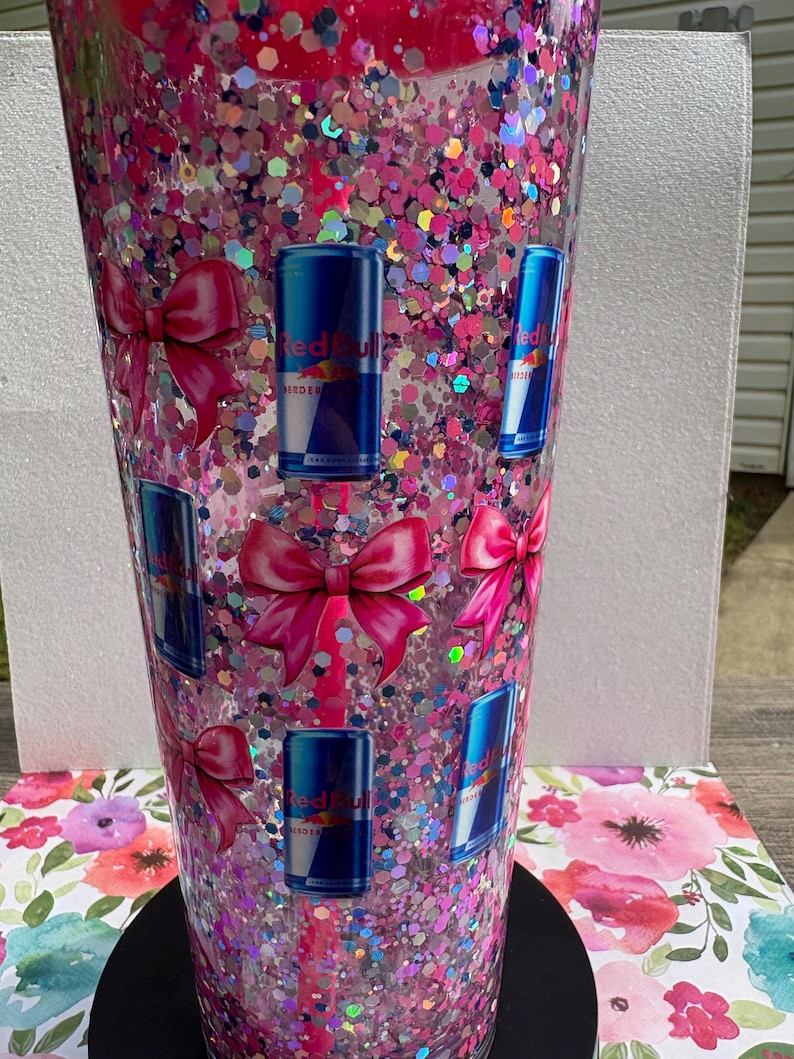May include: A tall, clear tumbler filled with pink glitter and Red Bull can decals. The tumbler also features pink bow decals. The tumbler sits on a black base and is set against a floral background.