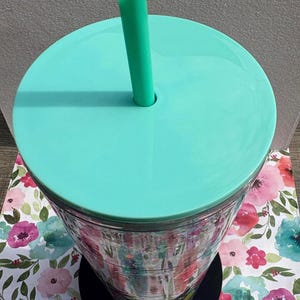 May include: A clear plastic tumbler with a vibrant floral pattern featuring pink, red, and teal watercolor-style flowers. The cup has a solid mint green lid and a matching mint green straw, with a black circular base at the bottom of the vessel.