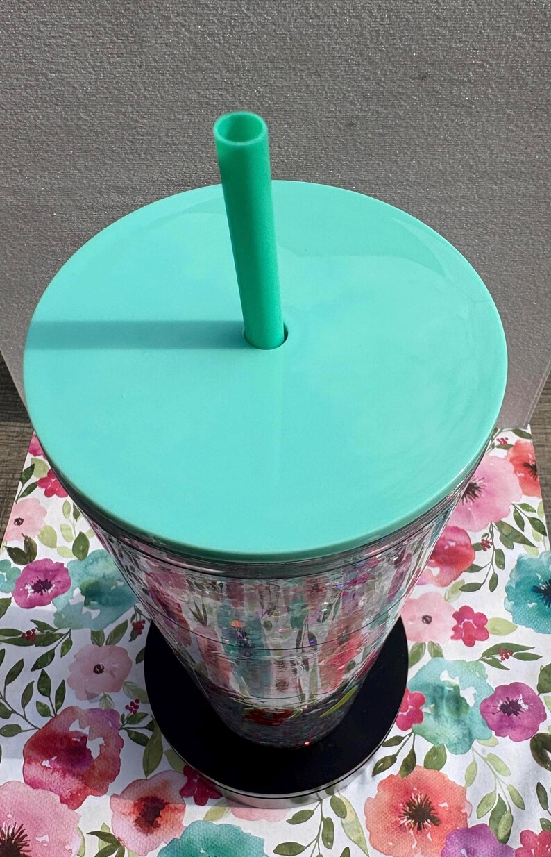 May include: A clear plastic tumbler with a vibrant floral pattern featuring pink, red, and teal watercolor-style flowers. The cup has a solid mint green lid and a matching mint green straw, with a black circular base at the bottom of the vessel.