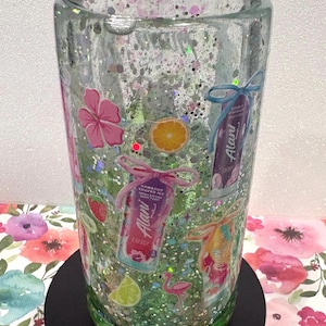 May include: A clear double-walled tumbler filled with green liquid and silver glitter. The exterior features decals of Alani energy drink cans, pink hibiscus flowers, orange and lime slices, strawberries, and a small pink flamingo graphic.
